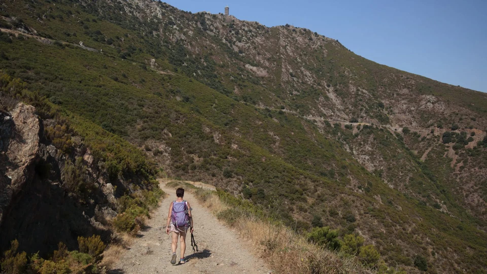 Hiker with backpack walking on dirt path below scrub-covered mountain with tower on summit.