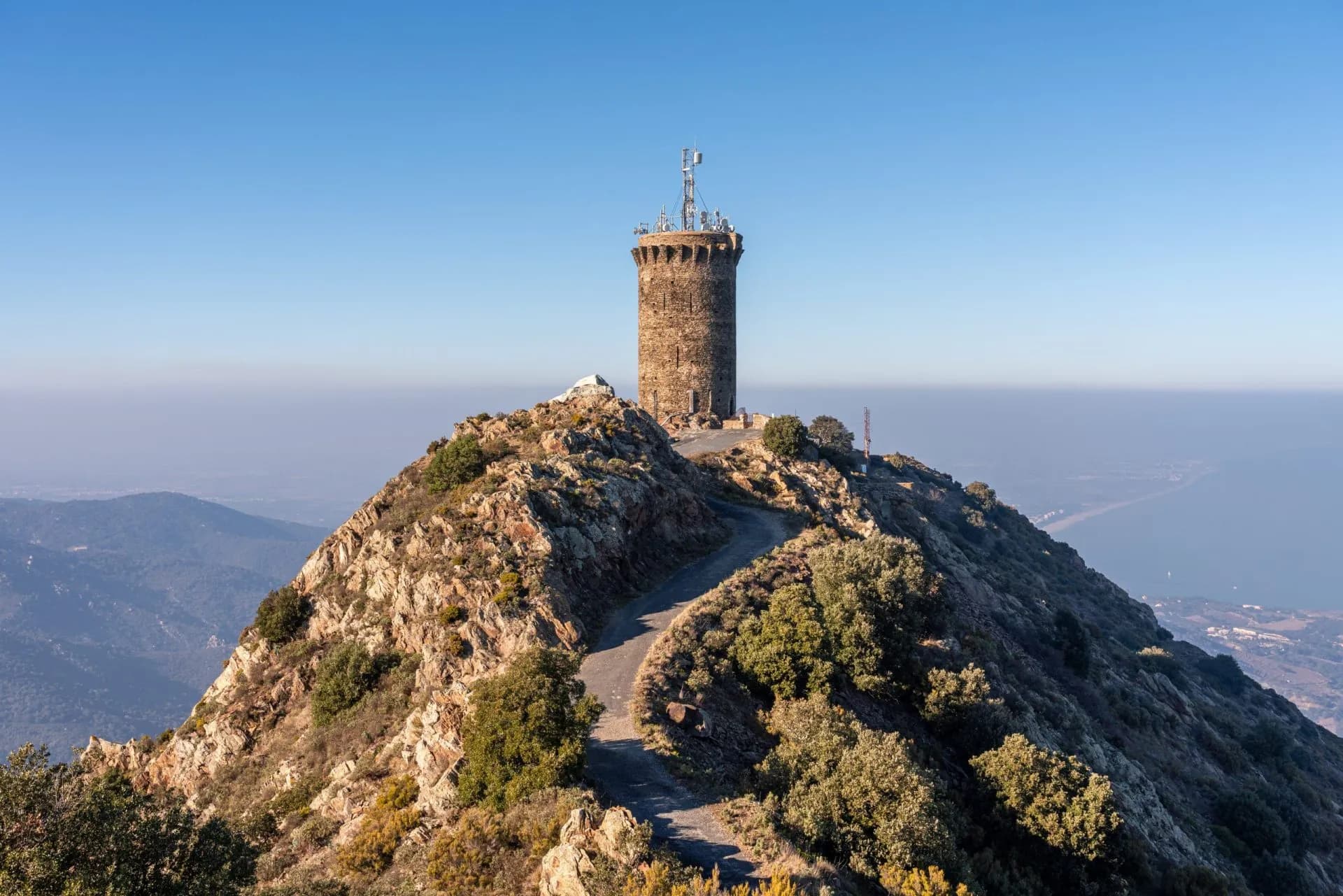 Stone tower with antennas on rocky peak overlooking hazy coastal landscape and sea