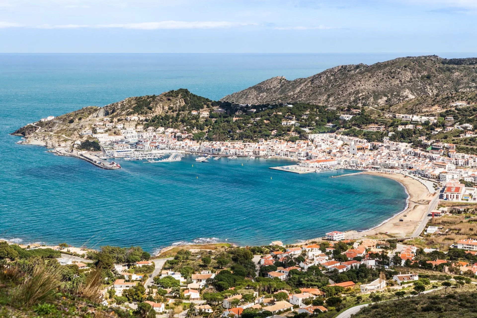 Coastal town of Port de la Selva with white buildings nestled against arid hillsides and a turquoise bay.