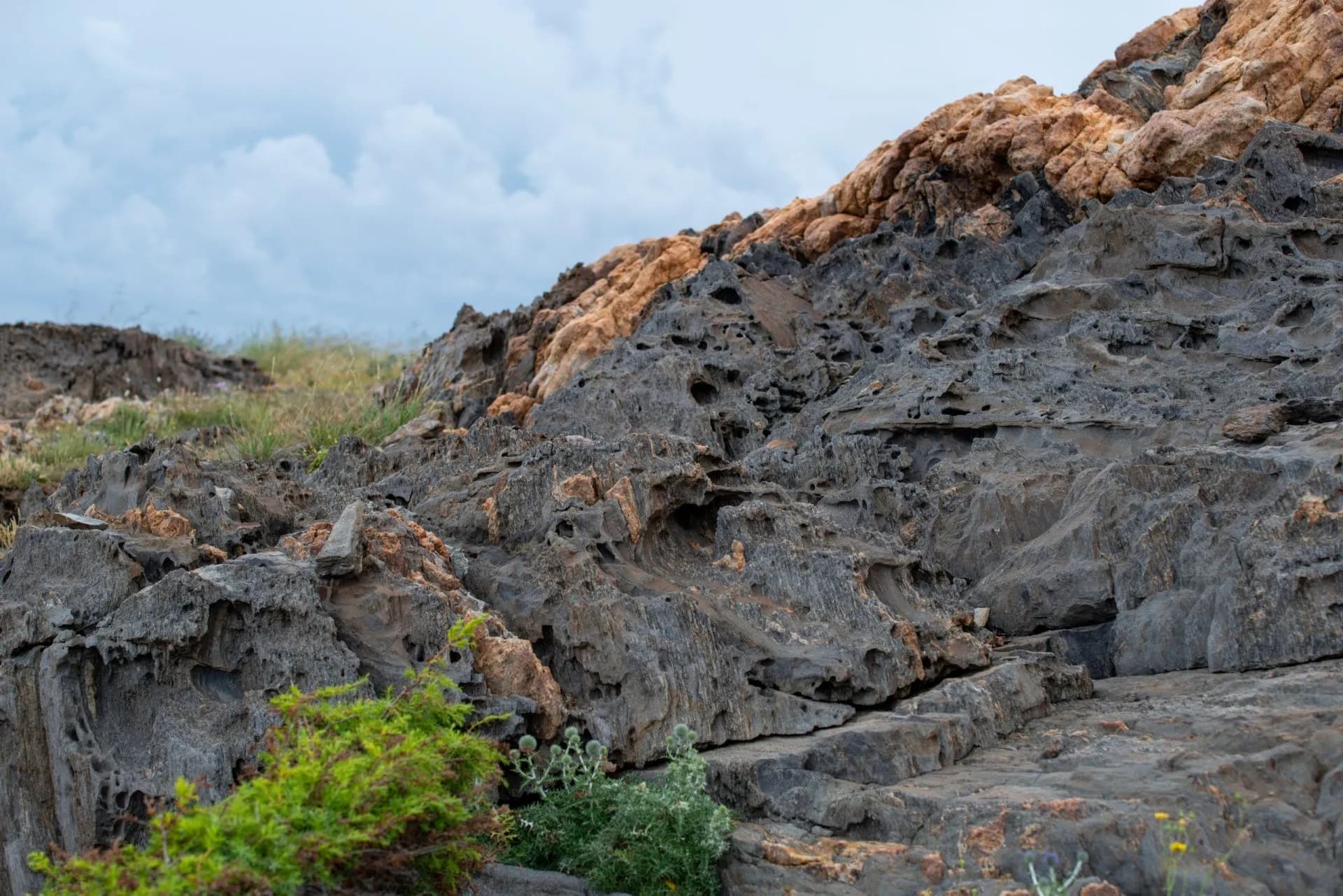 Eroded dark and orange rock formations with sparse green vegetation under a cloudy sky at Cap de Creus National Park.