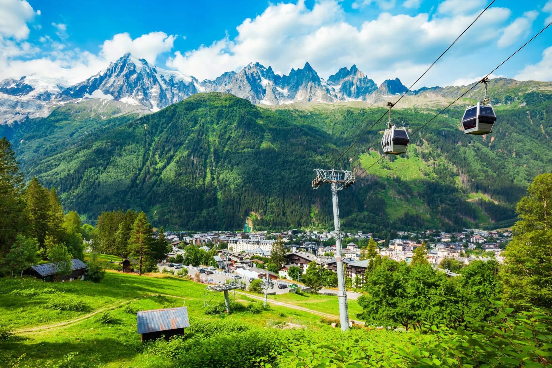Cable cars ascend over a green valley town with snow-capped alpine mountains in the background.