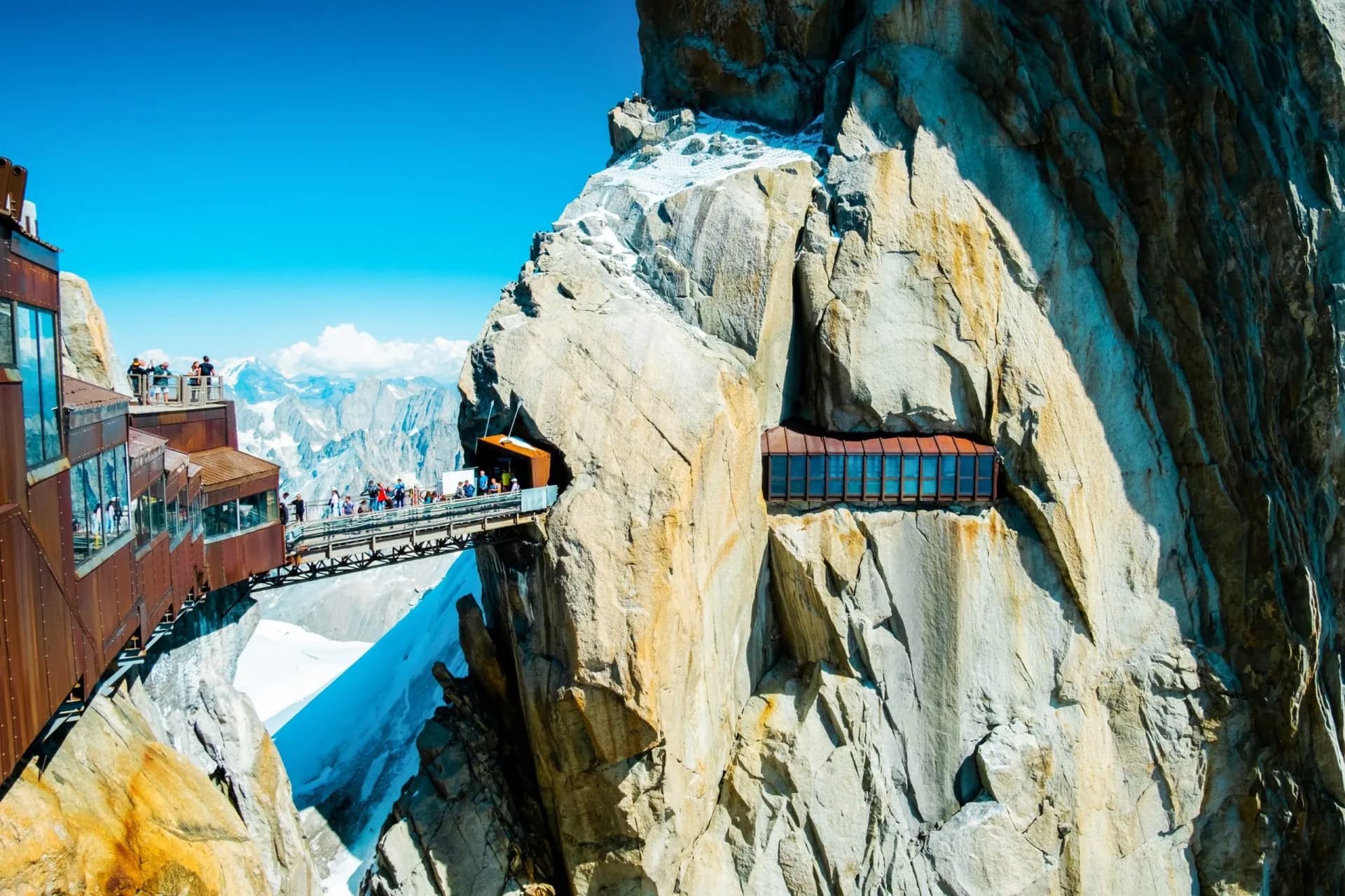 Footbridge connecting viewing platforms on a sheer rock face at Aiguille du Midi with snowy mountains.