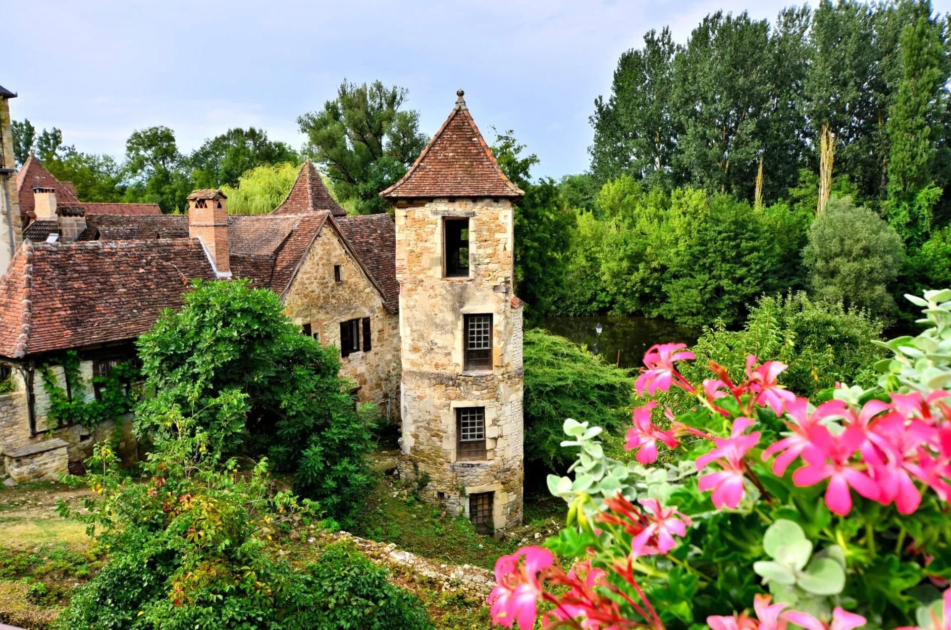 Stone village buildings with tile roofs surrounded by lush greenery and pink flowers in Carennac.