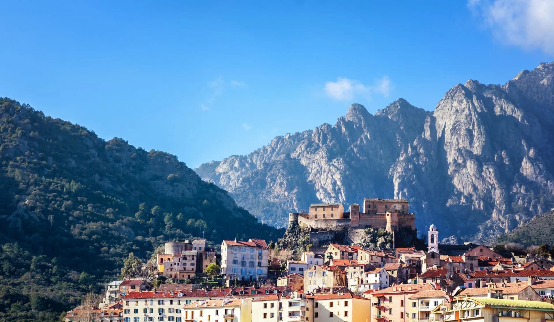 Hillside town with terracotta roofs below rugged mountains under a clear blue sky in Corsica.