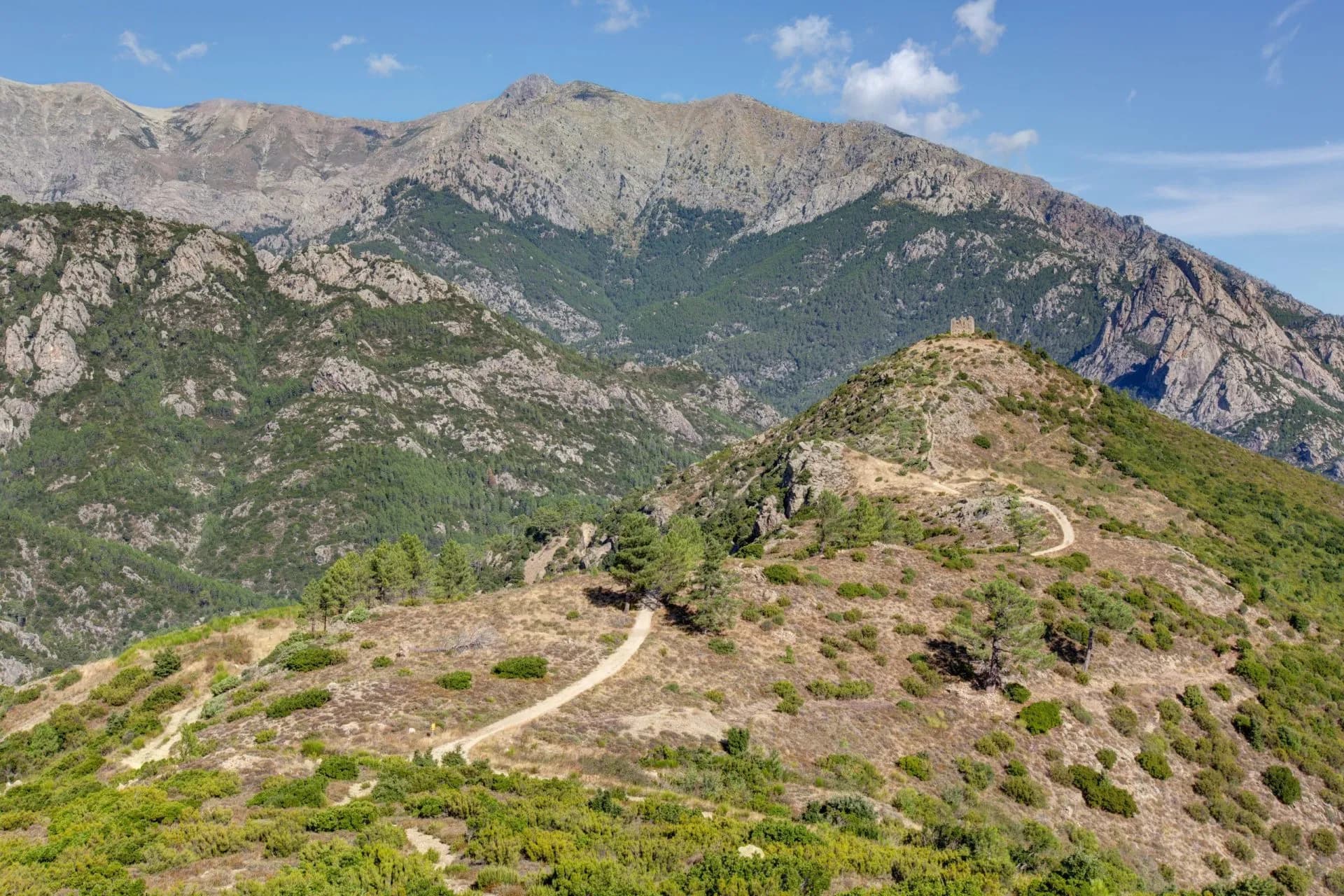 Hiking path winding up a dry, scrub-covered hill toward a small ruin with large mountains behind.