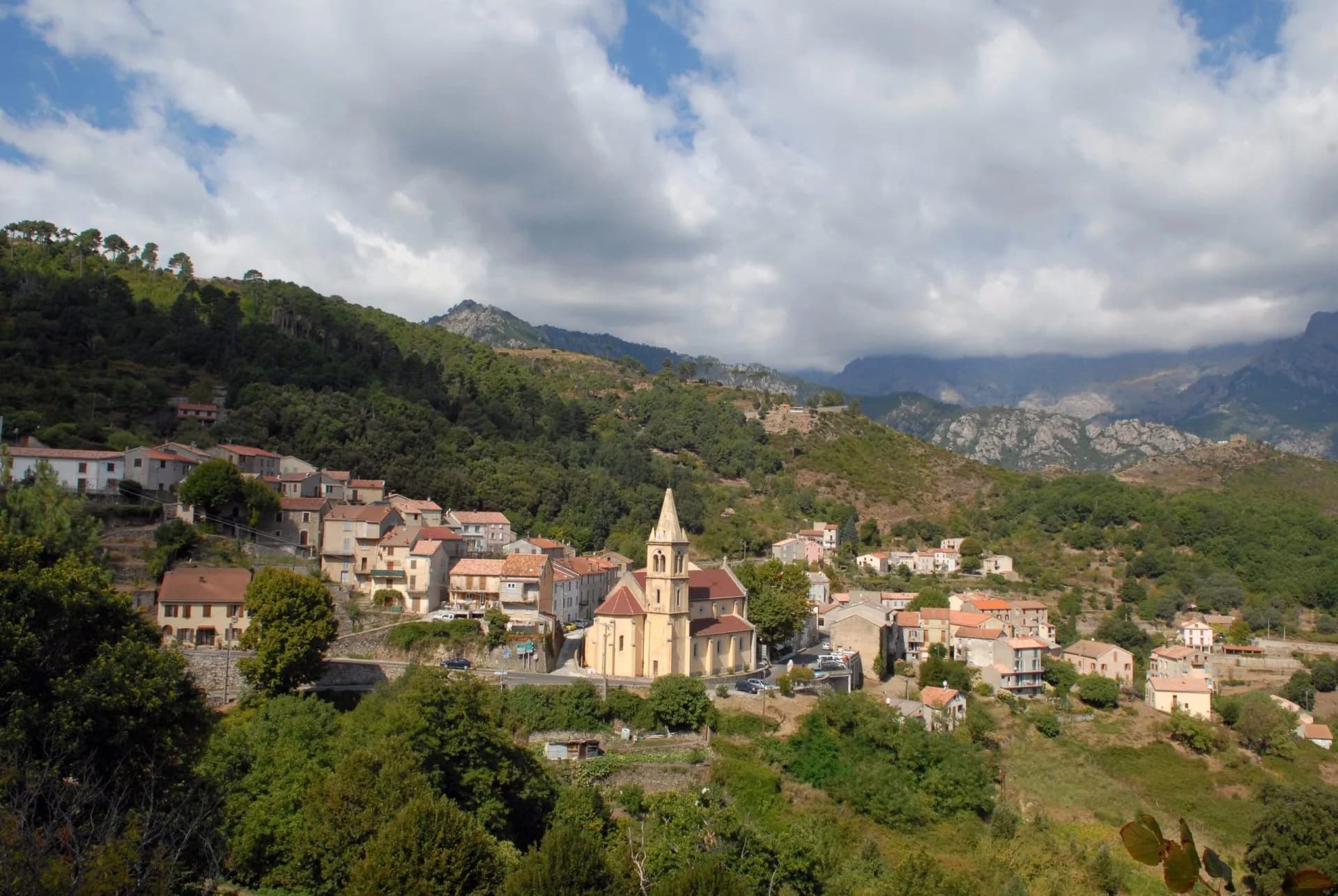 Hillside village with a prominent church nestled in lush green mountains under a cloudy sky, Vizzavona.