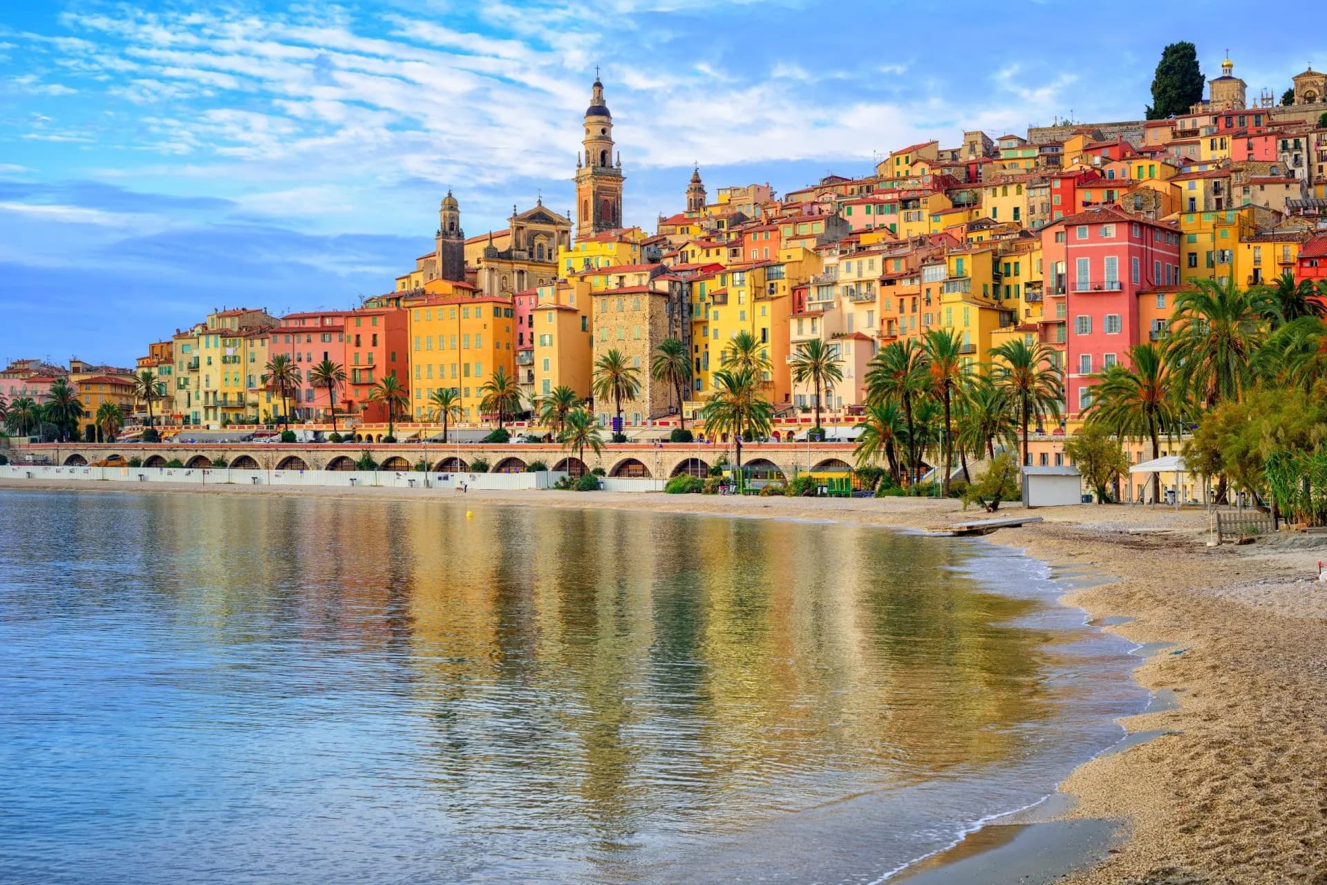 Colorful stacked buildings and palm trees line the beach in Menton reflected in the calm sea water.