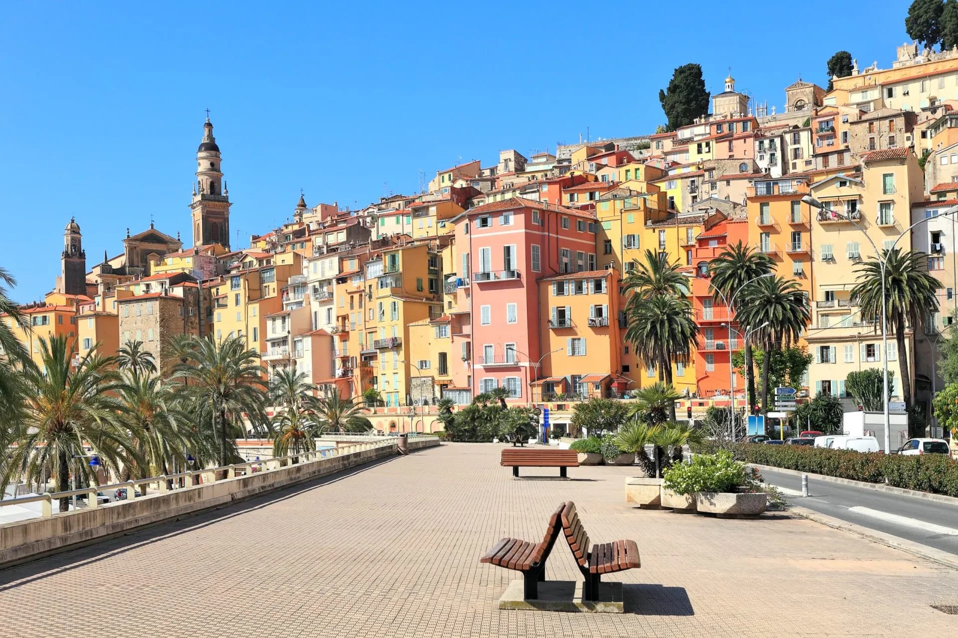 Promenade with benches facing colorful hillside buildings and palm trees in Menton.