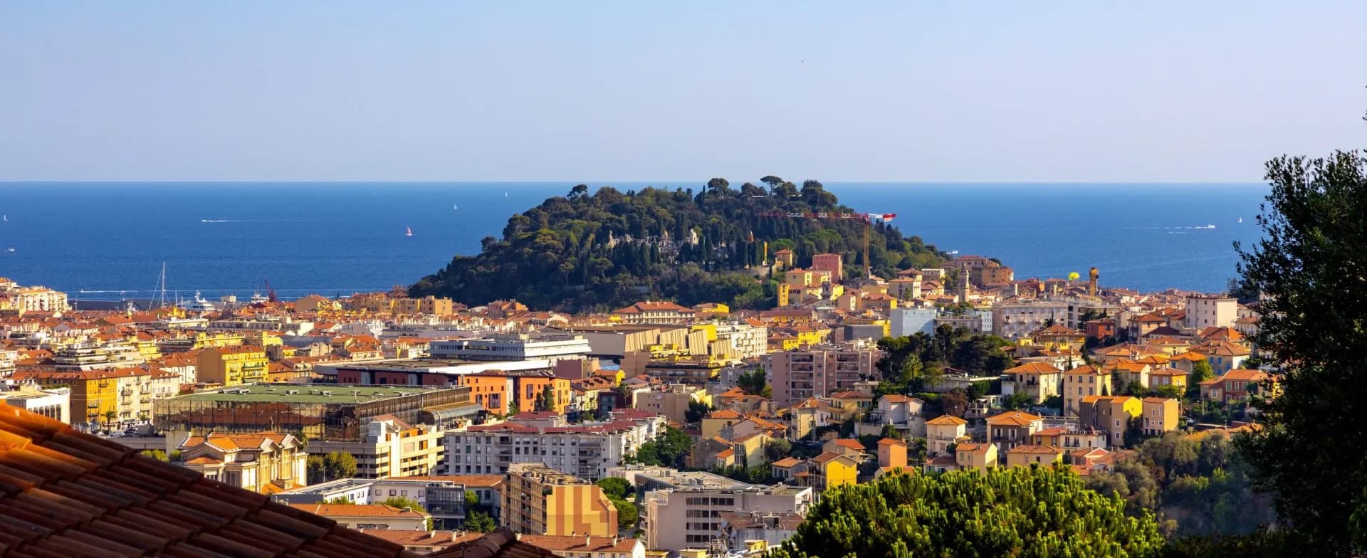Cityscape of colorful buildings leading to a wooded hill overlooking the deep blue Mediterranean Sea.