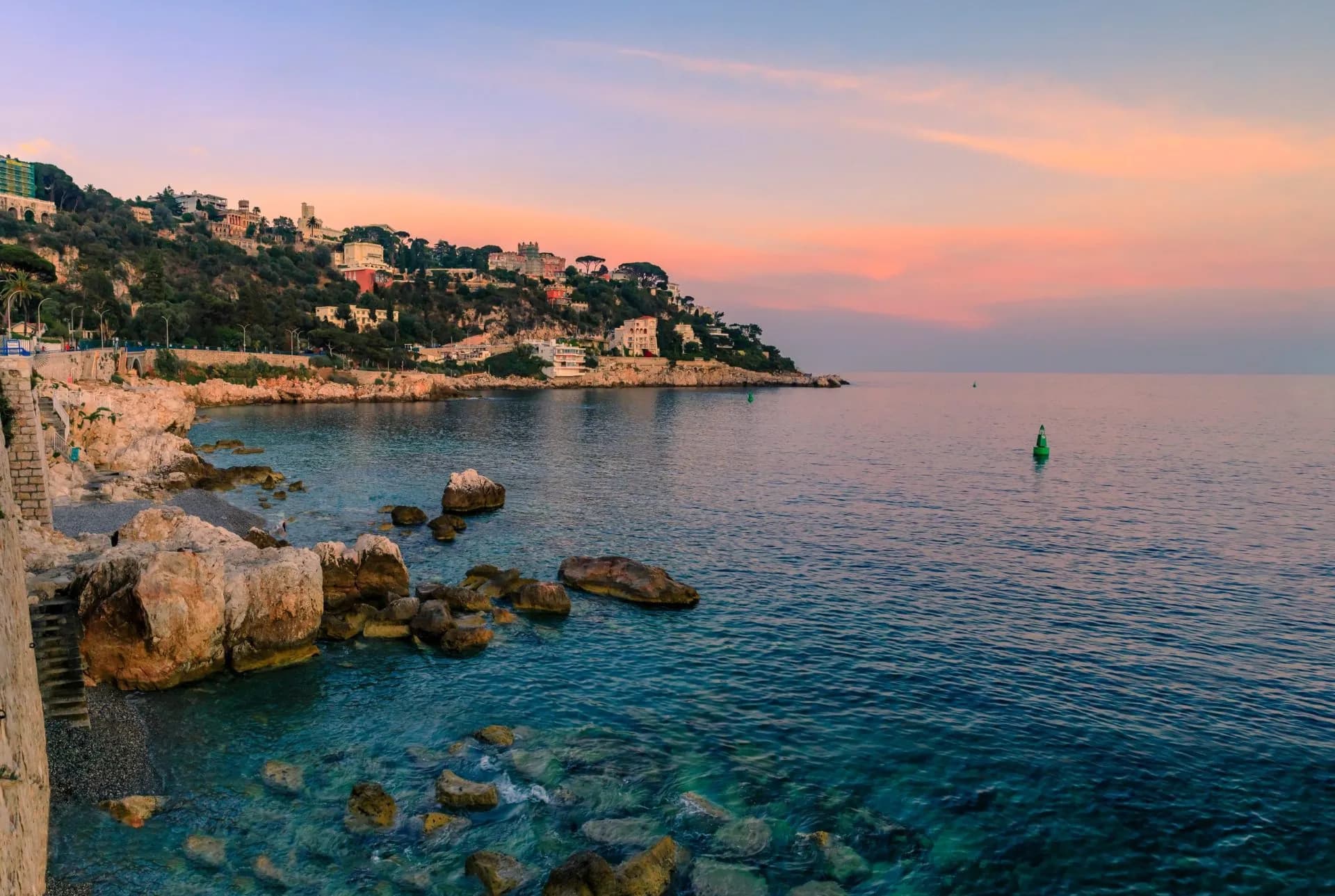 Rocky coastline and clear blue water near hillside buildings at sunset, near Mount Boron.