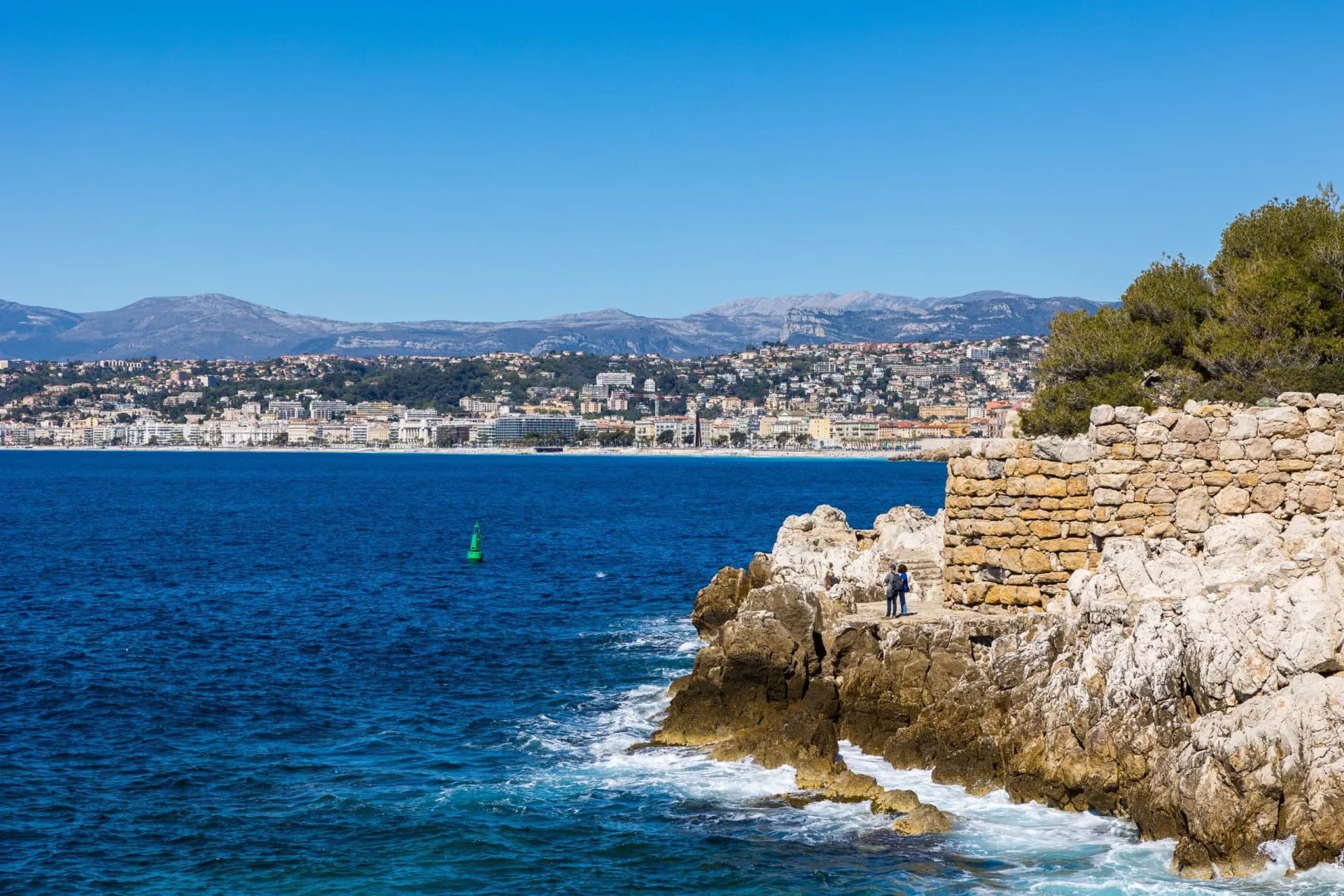 Rocky coastline with stone wall overlooking deep blue sea, Nice cityscape and mountains in background.
