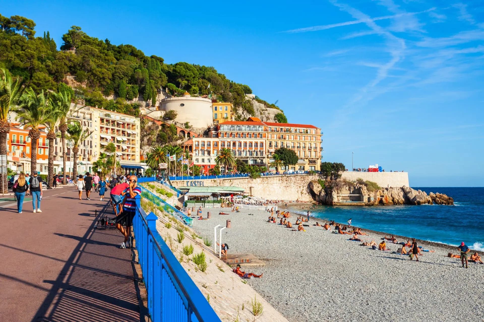 People walking by a pebble beach with blue water and buildings backed by a green hill in Nice.