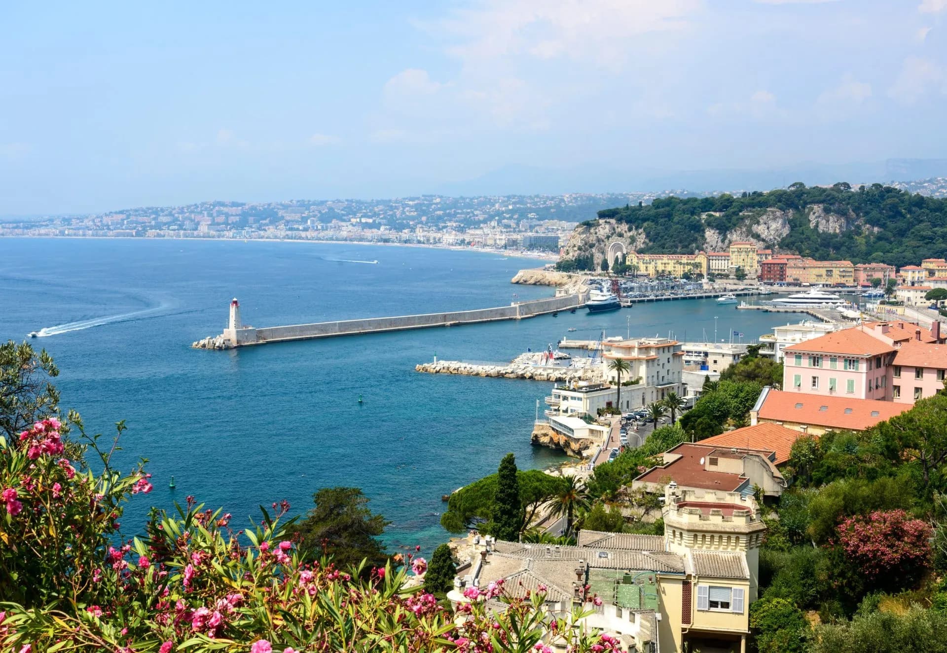 Coastal view of Nice harbor with lighthouse, colorful buildings, and pink flowers in foreground.