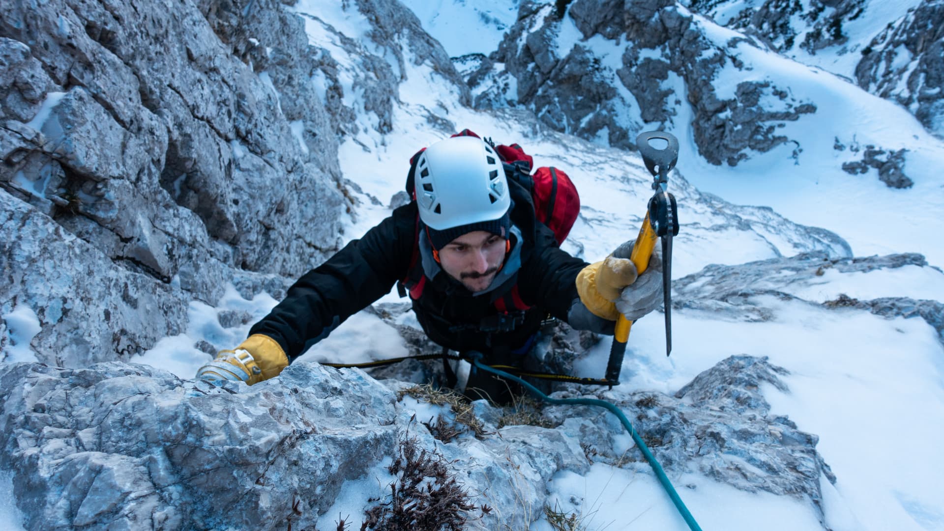 Winter mountaineer climbing icy rock face with ice axe in snowy alpine setting.