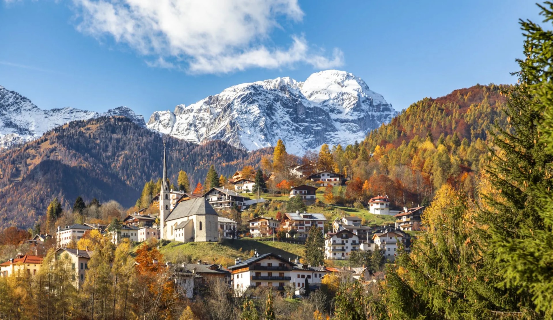 Alpine village Val di Zoldo with church nestled on hillside below snow-capped mountains in autumn.