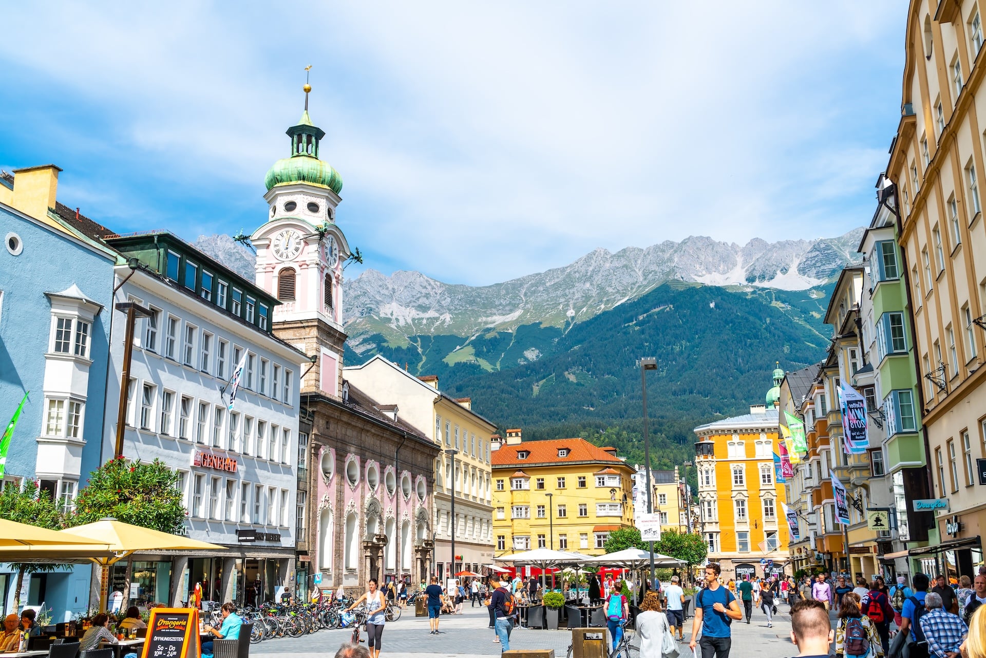 Crowded street with cafes and a clock tower in Innsbruck town center, backed by mountains.
