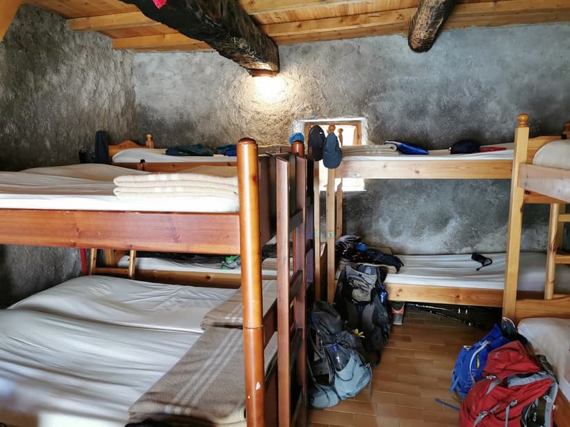 Bunk beds in a rustic room with stone walls, wooden beams, and hiking backpacks on the tiled floor.