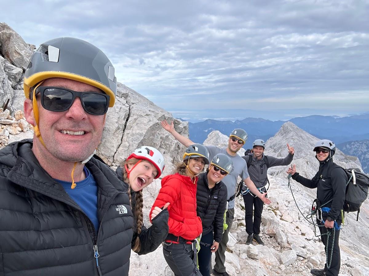 Group of hikers taking a selfie on a rocky mountain summit with distant peaks under cloudy sky.