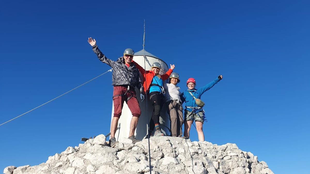 Four hikers with helmets celebrate on a rocky mountain summit next to a small white structure.