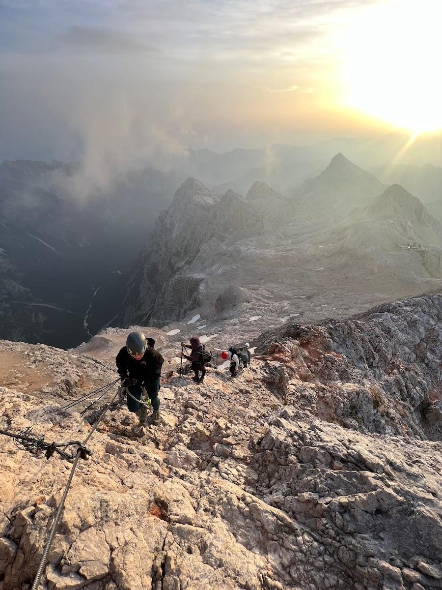 Hikers using via ferrata cables on rocky mountain ridge at sunrise with sun flare