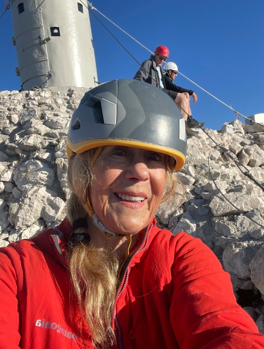 Woman in helmet takes selfie on rocky mountain summit near tower structure.