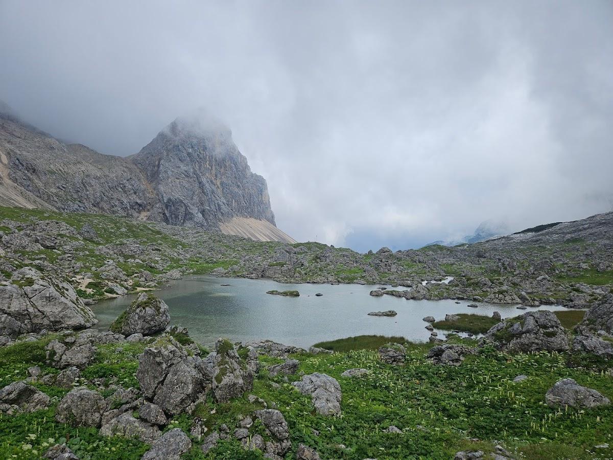 Alpine lake surrounded by large rocks, green vegetation, and a steep mountain peak shrouded in fog.