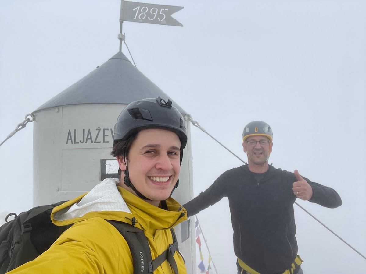 Two hikers with helmets at the Aljažev Stolp summit marker in heavy fog.
