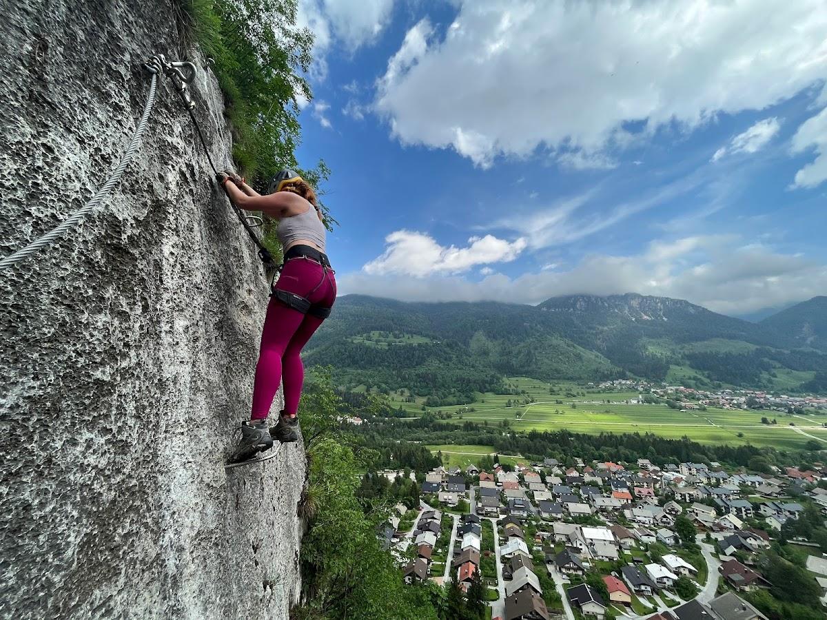 Woman climbing rock face with via ferrata cables overlooking green valley and town.