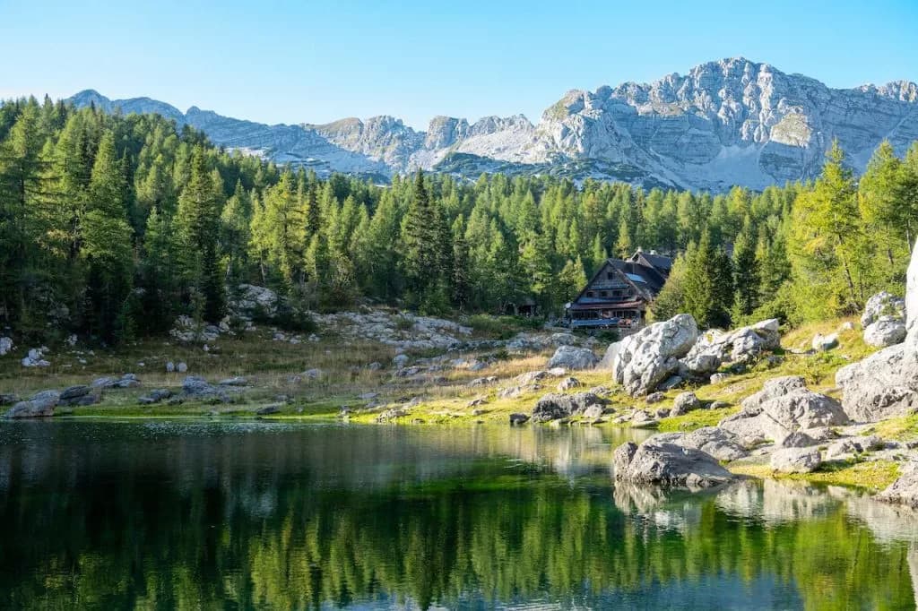 Mountain hut by Triglav Lakes reflecting in dark water with rocky shore and pine forest.