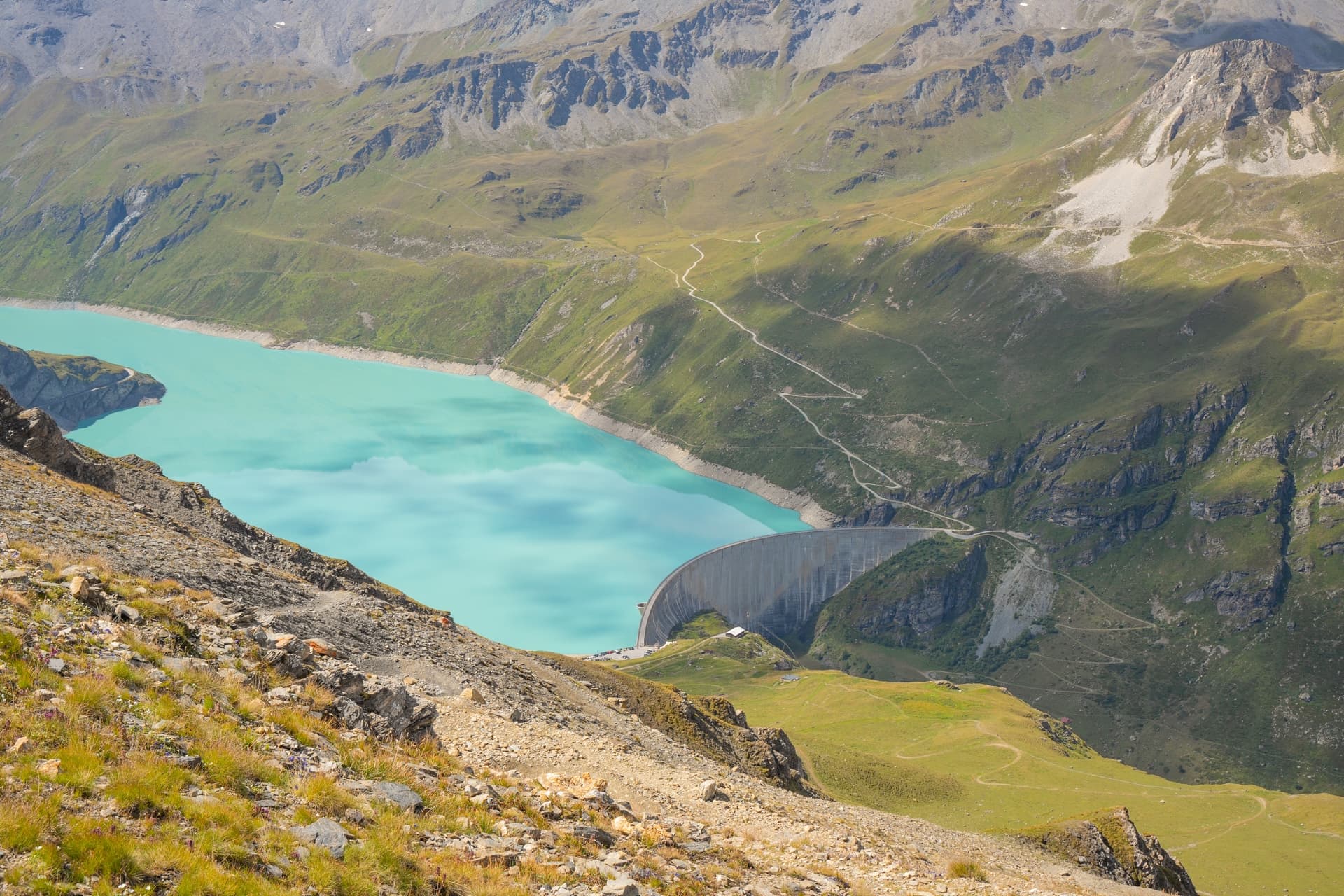 High-angle view of a large curved dam holding back turquoise reservoir water in a steep green mountain valley.