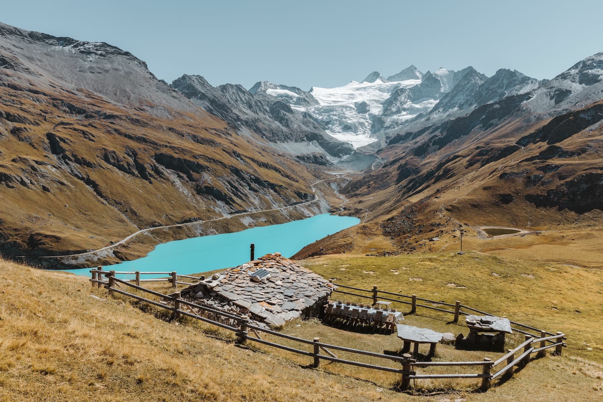 Stone hut with slate roof near turquoise alpine lake and glacier-capped mountains.