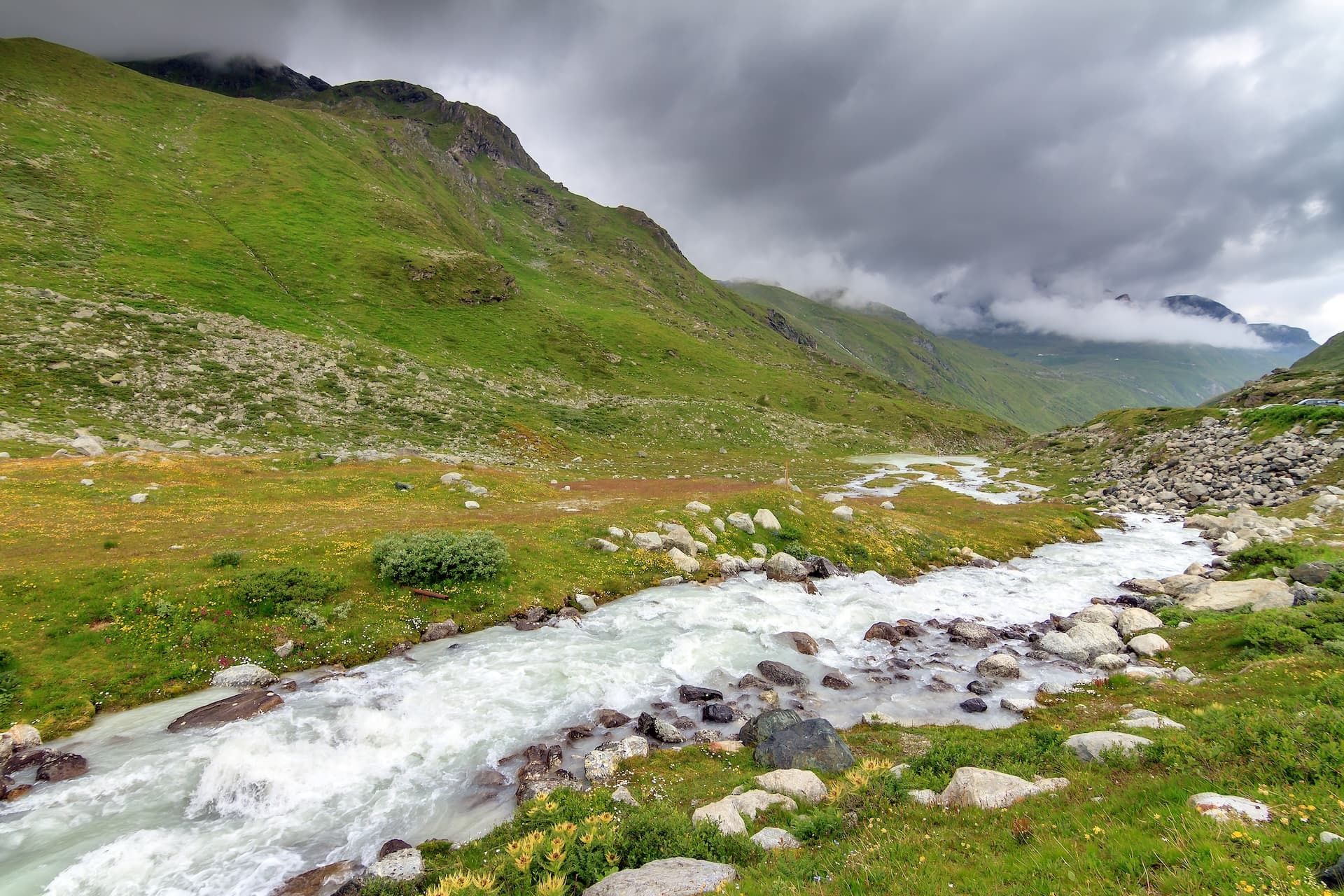 Rushing river through a green alpine valley under dark, cloudy skies in Valais.