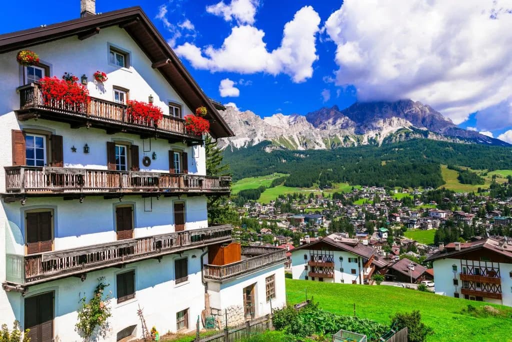 Alpine village with white houses and balconies below rugged, forested mountains under blue sky.