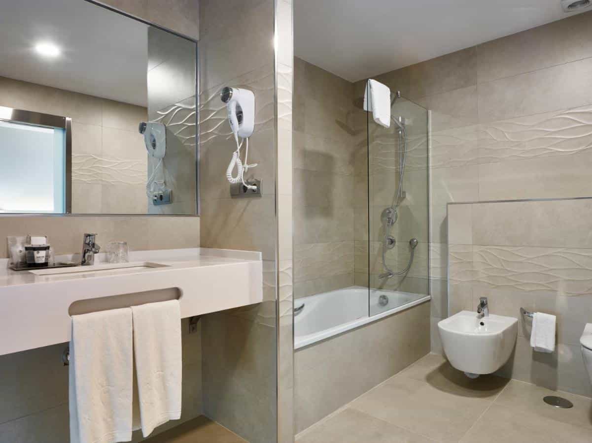 Hotel Maestrana modern bathroom with white vanity, bathtub, and bidet in beige tile.