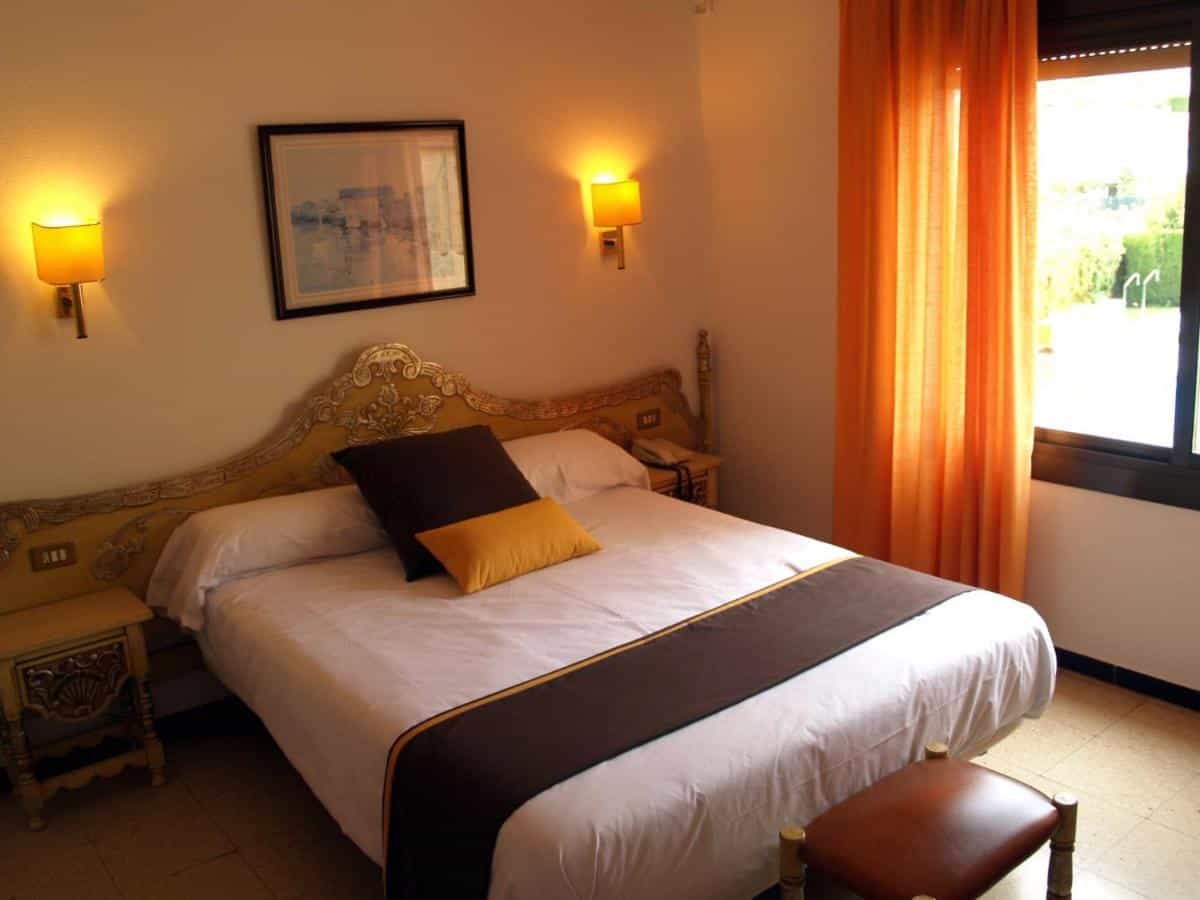 Hotel room with ornate yellow headboard, white bedding, and bright orange curtains.