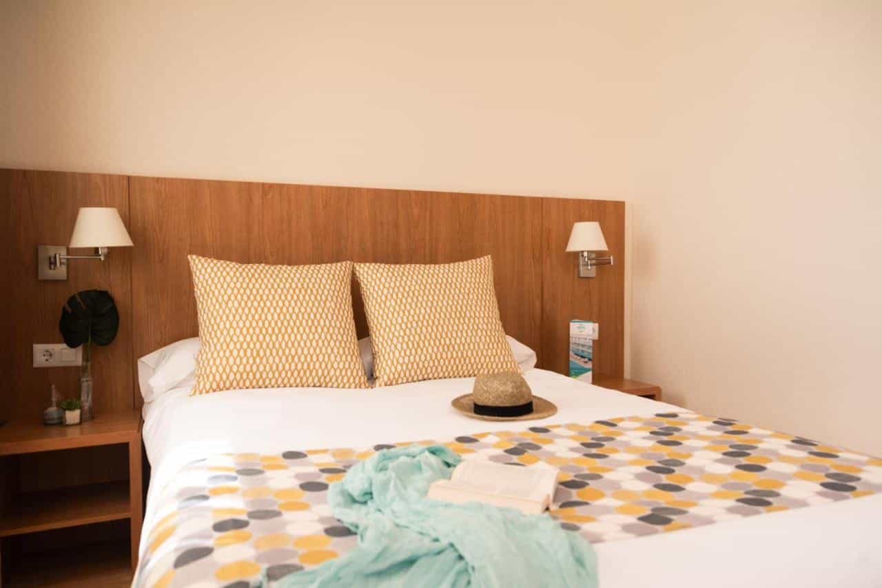 Aparthotel Ona Palamós room with a double bed, yellow pillows, patterned throw, and straw hat.