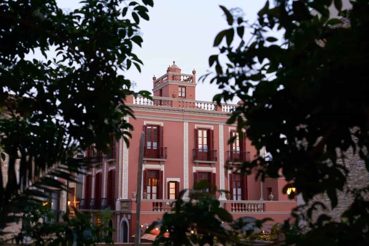 Pink historic building, Hotel Aiguaclara, framed by dark green foliage at dusk