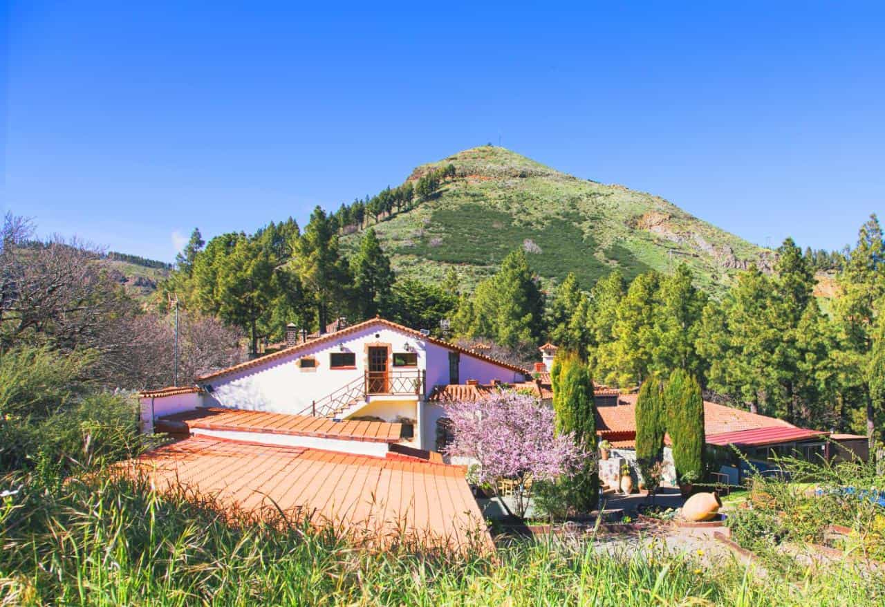 Rural hotel building with terracotta roof set against a steep, green mountain under a clear blue sky.