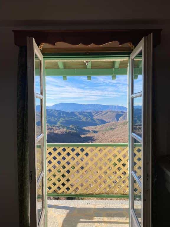 Mountain view from open balcony doors over forested hills under a blue, cloudy sky.