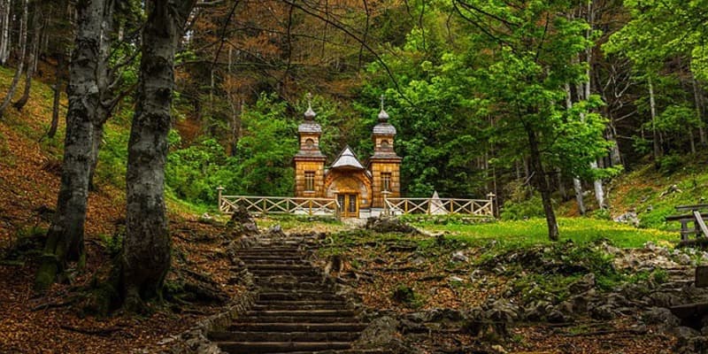 Wooden Russian chapel nestled in a dense forest with stone steps leading up to it.