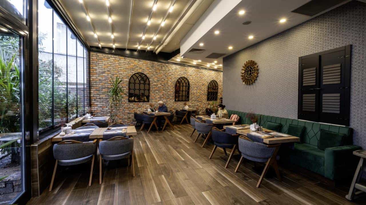 Restaurant dining area with brick wall, green velvet seating, and large windows overlooking greenery.