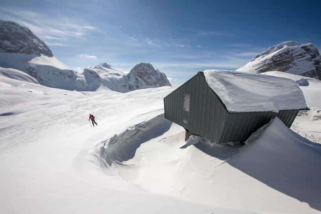 Skier descending snowy slope near modern mountain bivouac hut under blue sky.
