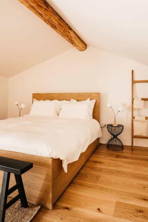 Bedroom with wooden bed, white linens, exposed beam, and ladder shelf in attic space.