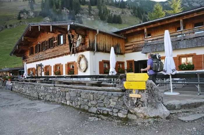 Hiker with backpack arriving at wooden alpine hut with stone wall and yellow trail signs