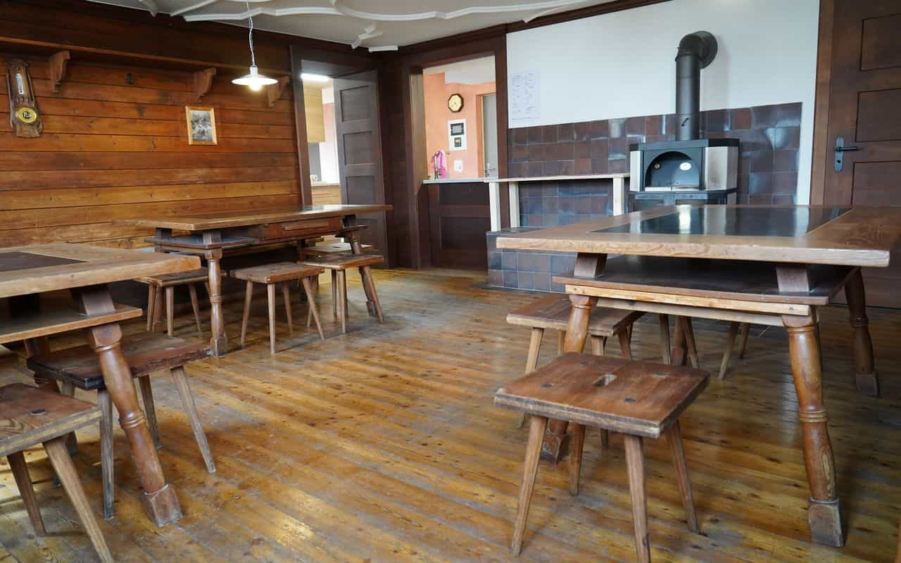 Rustic dining room with wood paneling, heavy wooden tables, stools, and a wood-burning stove.