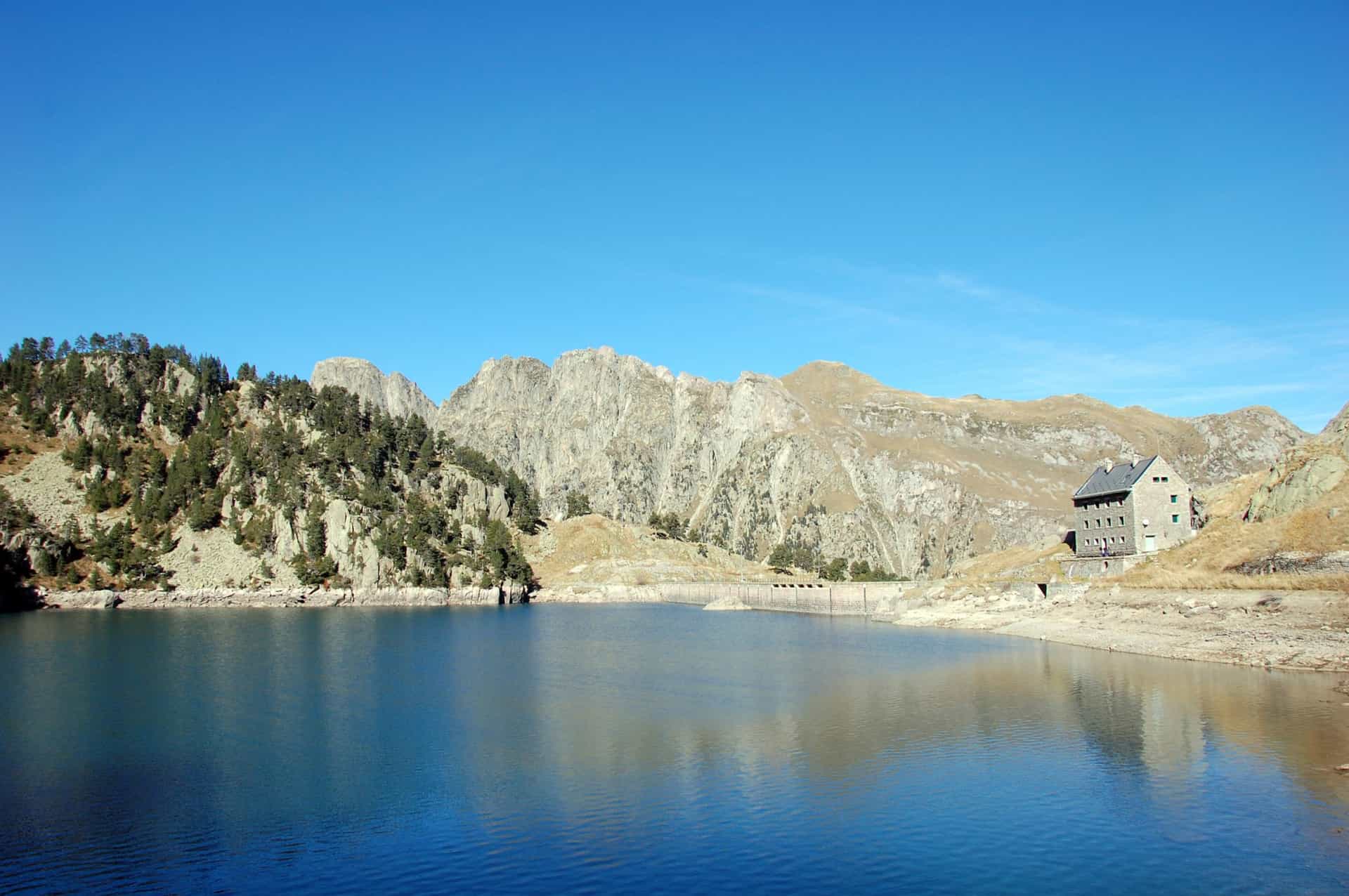 Mountain refuge by reservoir with low water level, rocky slopes, and clear blue sky.