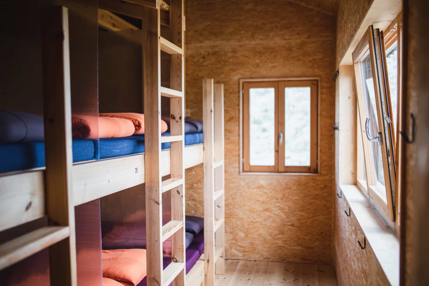 Wooden bunk beds with orange and blue bedding inside a room with OSB walls and wooden windows.