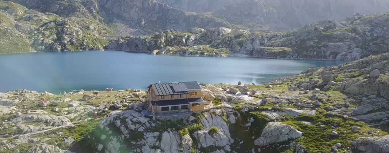 Mountain refuge with solar panels beside a blue alpine lake surrounded by rocky terrain.