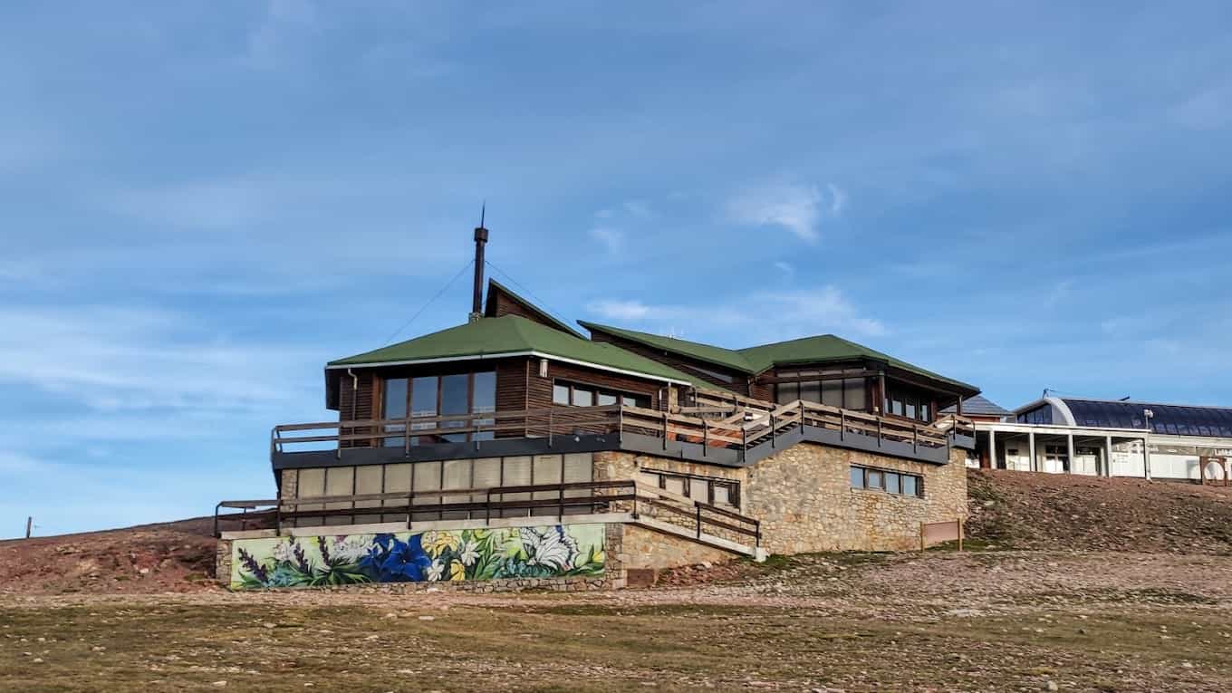 Mountain lodge with green roof and wooden balconies on grassy hillside under blue sky.