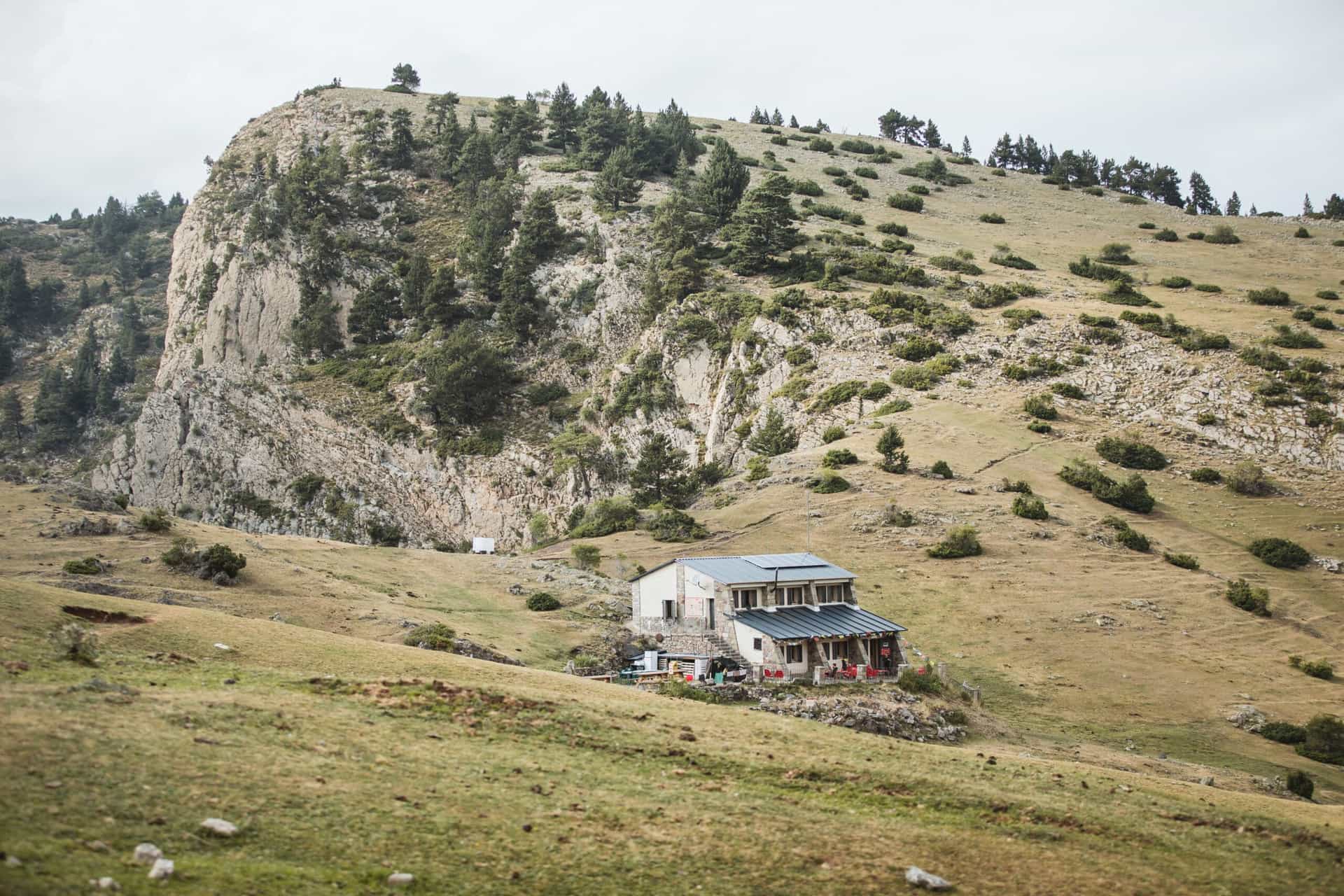 Mountain refuge Refugi Prat d'Aguiló nestled against a large rocky cliff face with sparse vegetation.